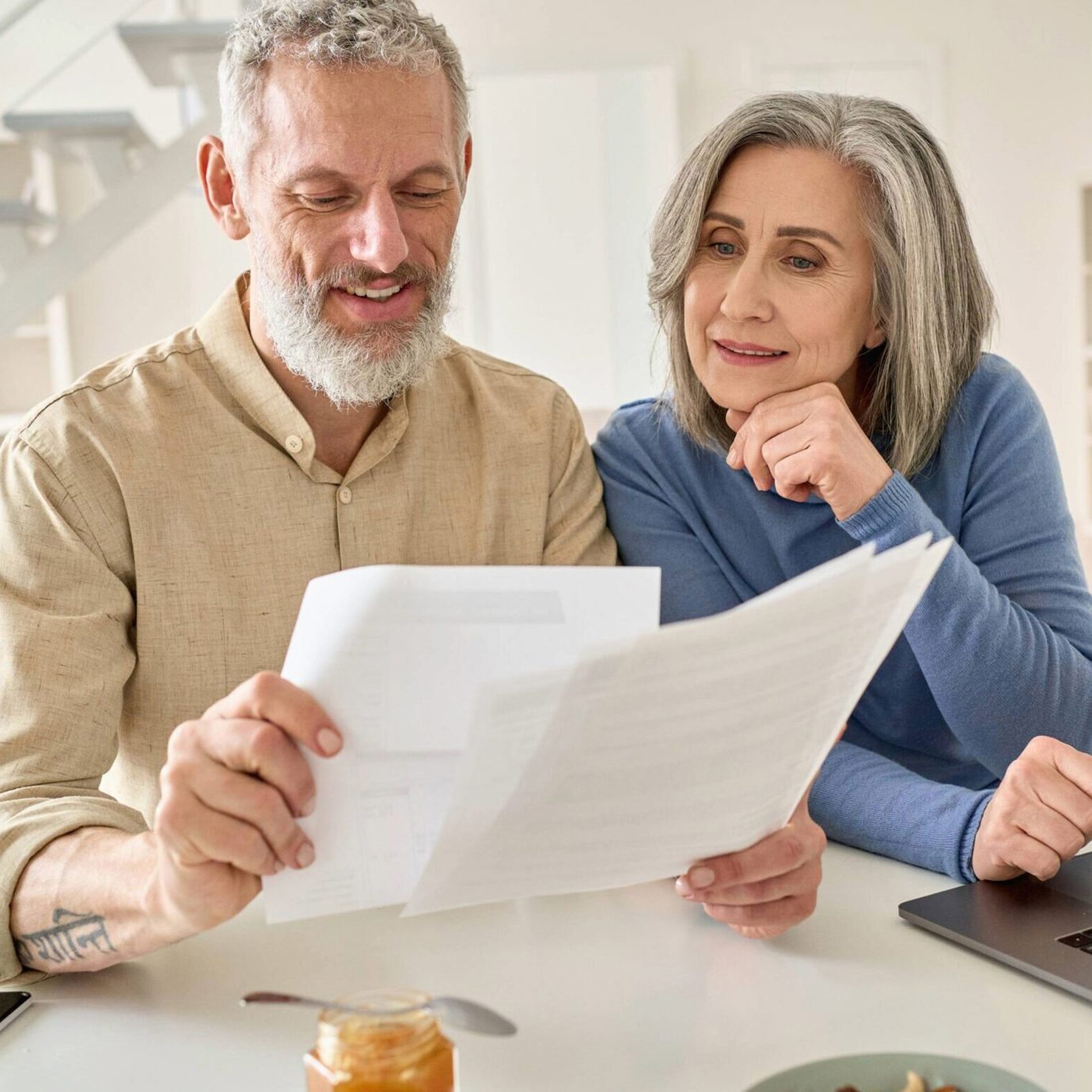 Smiling senior couple sitting together at home reviewing paperwork beside a laptop.