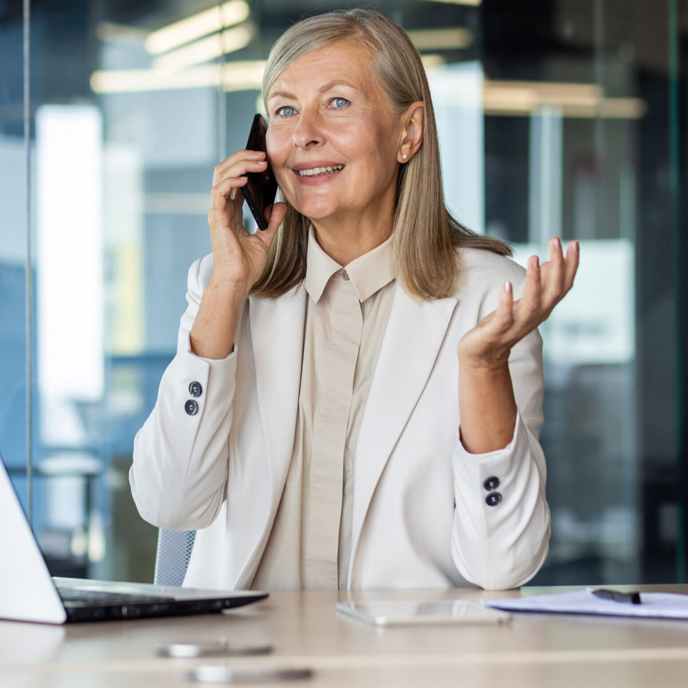Smiling woman talking on a mobile phone while sitting at a desk with a laptop in an office.