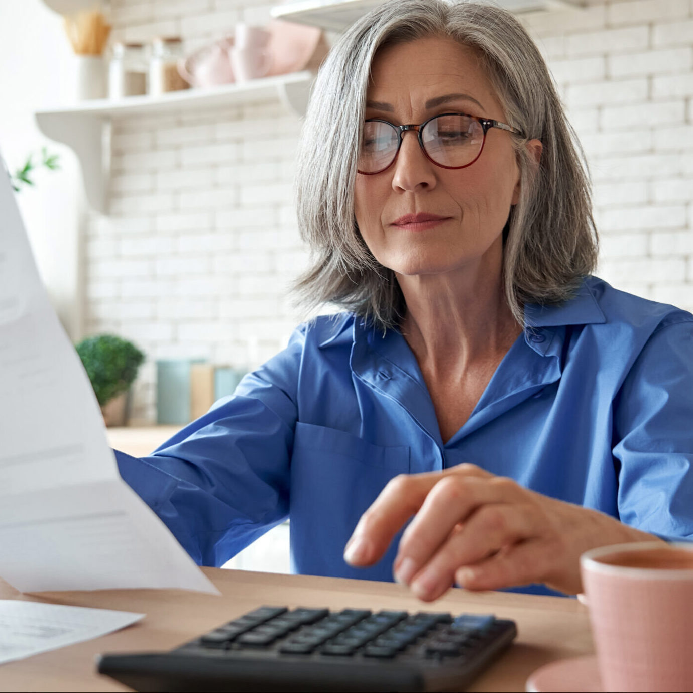 Senior woman with glasses reviewing paperwork while sitting at a table at home.