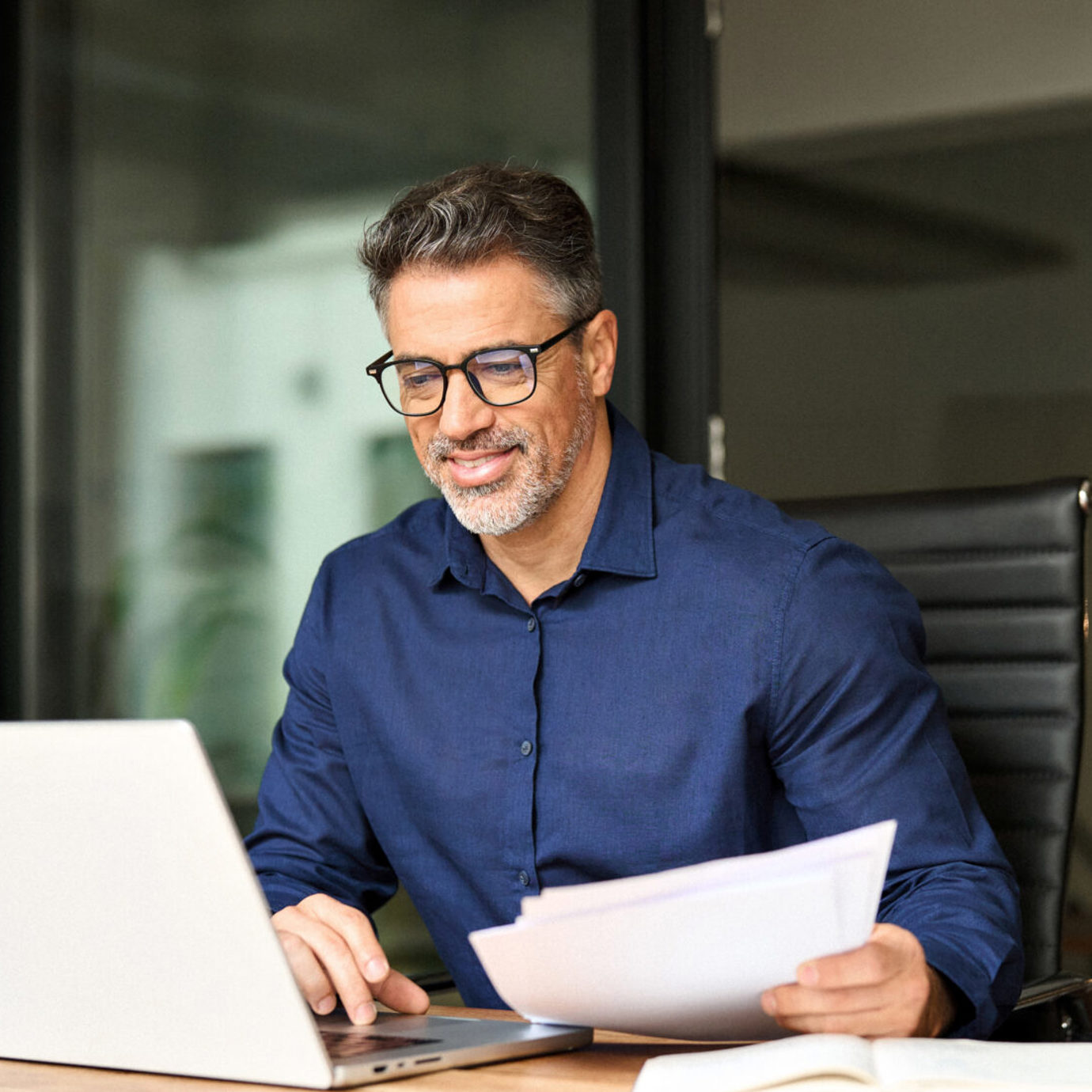 Smiling middle-aged man wearing glasses reviewing paperwork while working on a laptop at an office desk.