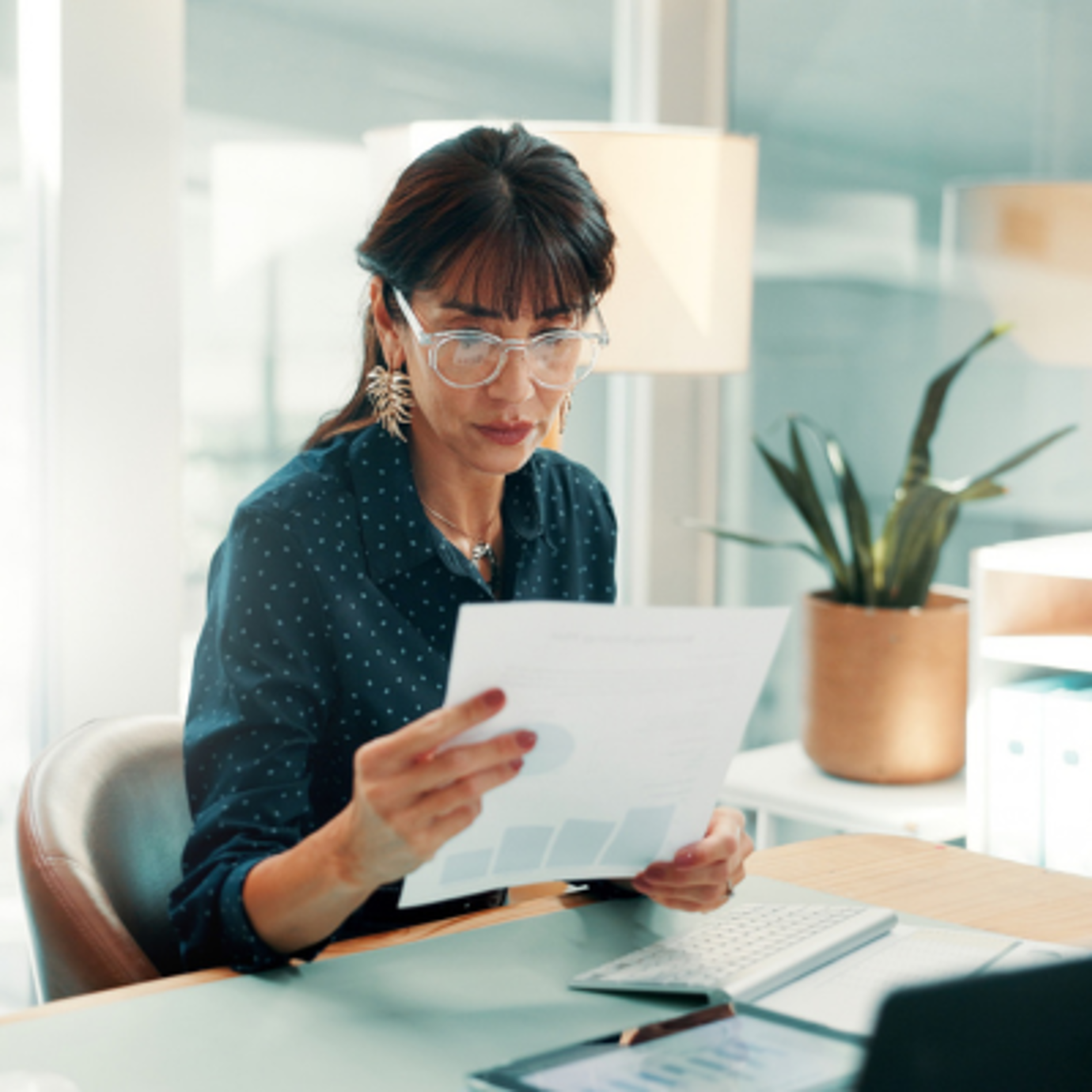 Woman wearing glasses reviewing paperwork while sitting at a desk at home.