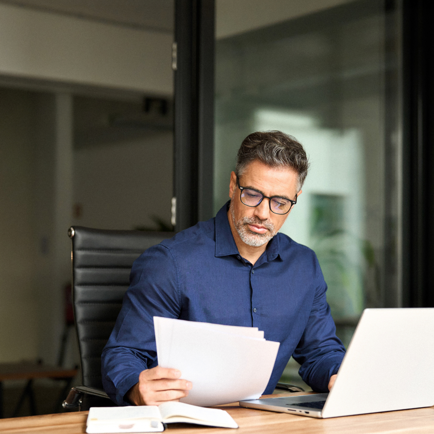 Man wearing glasses reviewing paperwork while working on a laptop at an office desk.