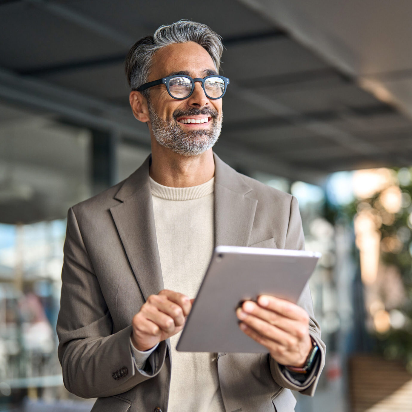 Smiling  man wearing glasses and a blazer while holding a tablet.